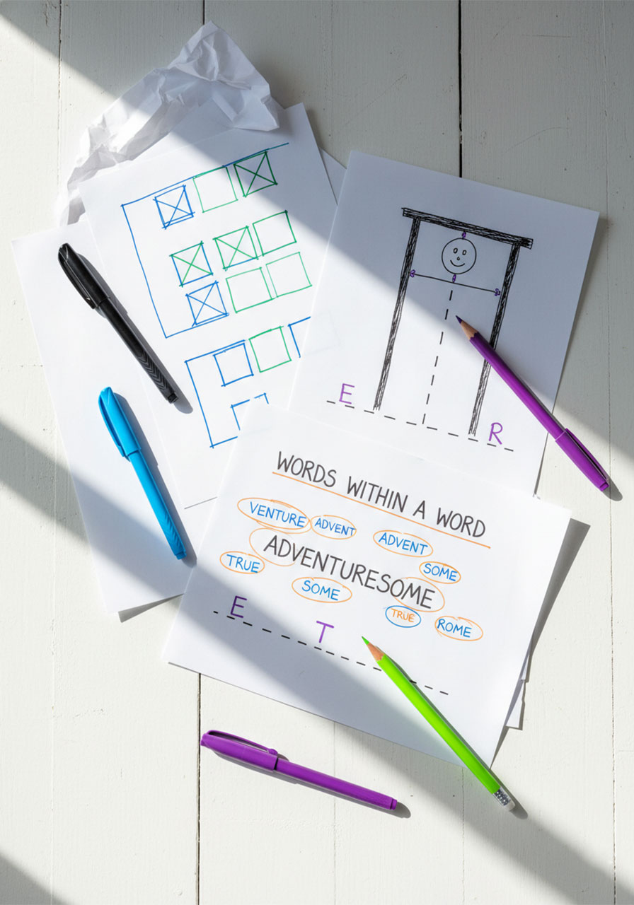 Overhead flat lay of multiple fun pen and paper games including dots and boxes and hangman on a white table with colorful pens
